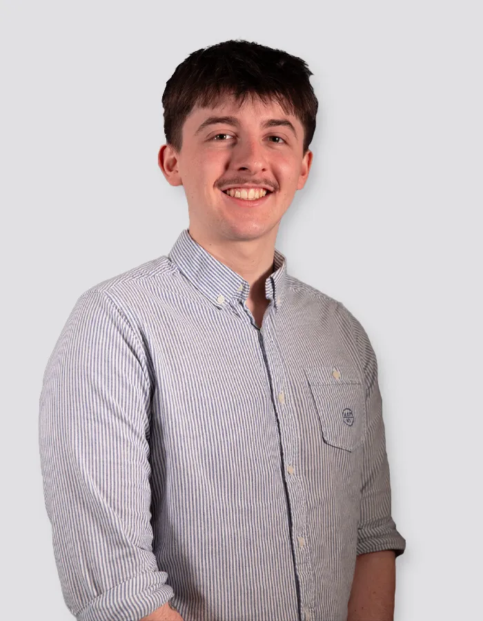 Young man with short brown hair and mustache wearing a light blue and white striped button-up shirt, smiling against a plain light gray background.