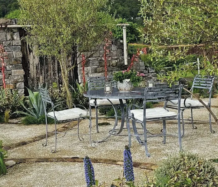 Outdoor garden seating area with metal table and four cushioned chairs surrounded by plants and stone walls.