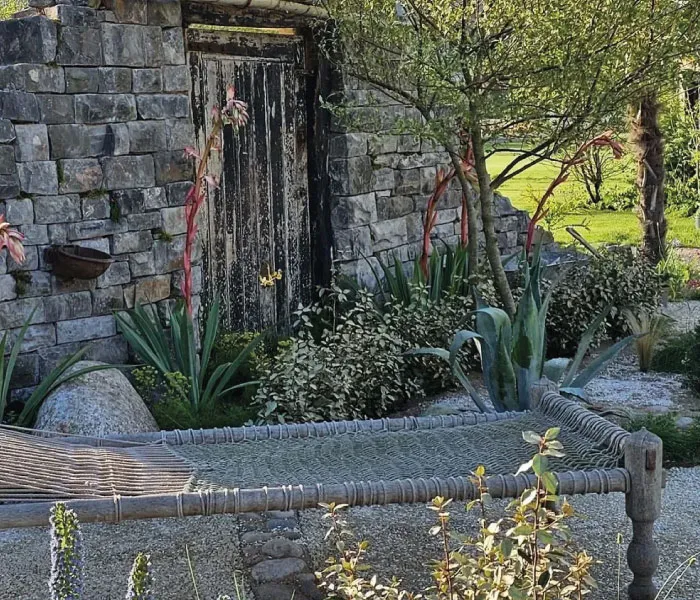 Rustic stone wall with weathered wooden door surrounded by lush garden plants and a woven outdoor daybed.