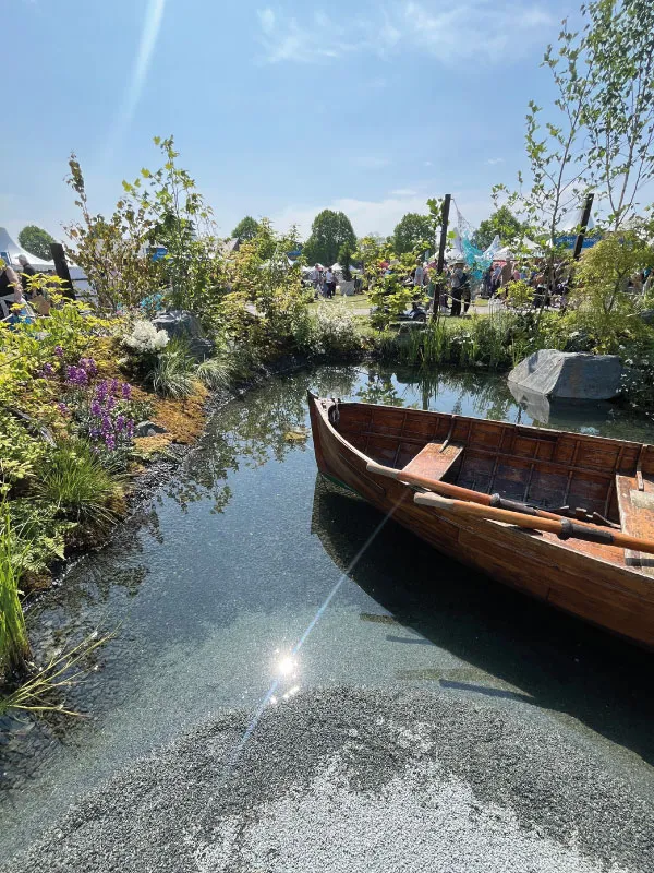 Wooden rowboat on clear water surrounded by plants and people at an outdoor garden event.
