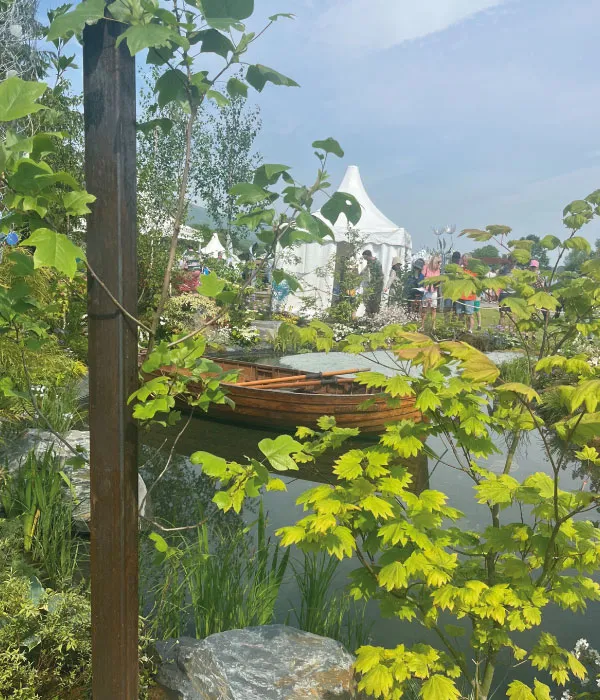 Wooden boat on calm water surrounded by green plants with event tents and people in the background.