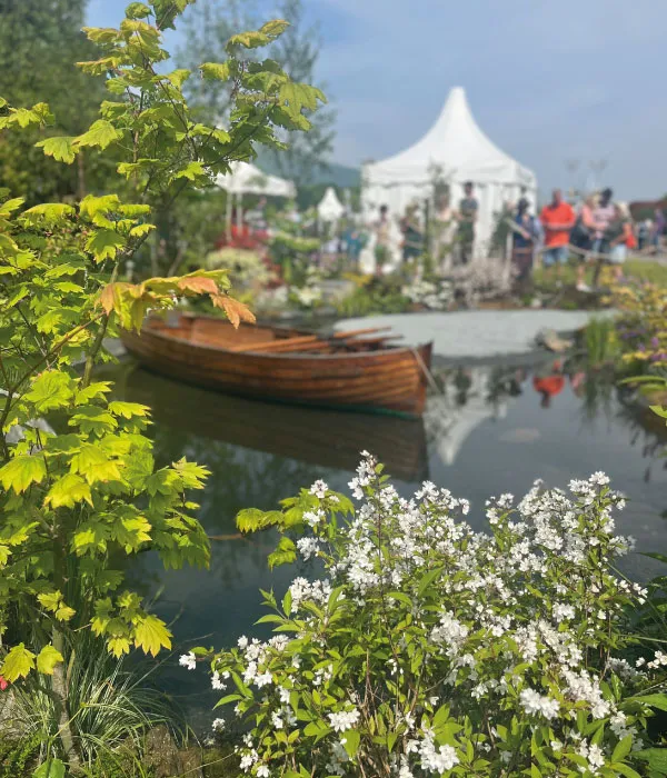 Wooden boat on a calm pond surrounded by green and flowering plants with people near white tents in the background.