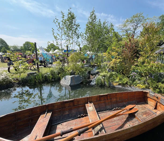 Wooden rowboat with two oars on calm pond surrounded by greenery and people in the background on a sunny day.