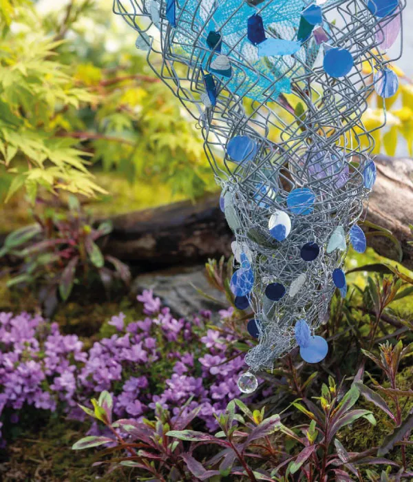 Blue and purple stained glass pieces attached to wire cone sculpture over garden with purple flowers and green foliage.