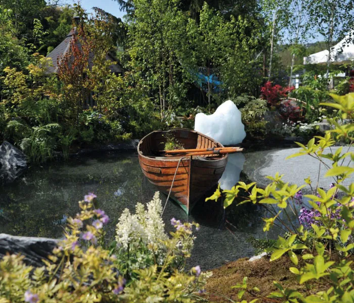 Wooden rowboat floating on a calm pond surrounded by green trees and colorful flowers.