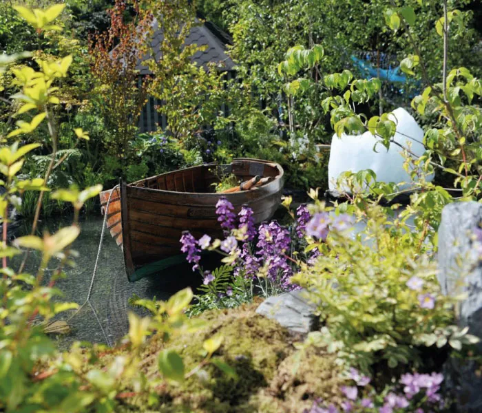 Wooden rowboat moored in a lush garden pond surrounded by green foliage and purple flowers.