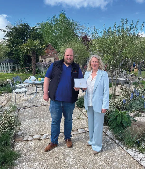 Man and woman standing on a stone path in a garden holding a certificate on a sunny day.