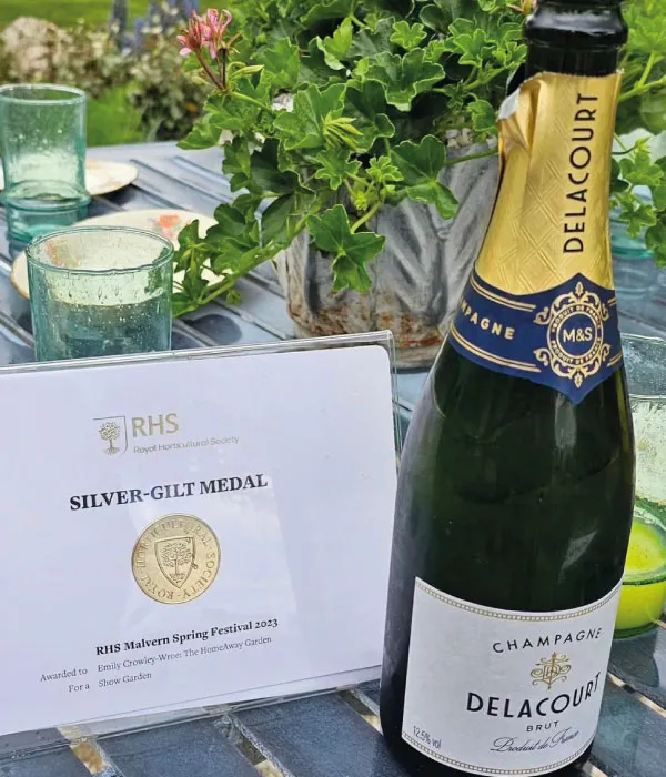 Bottle of Delacourt Champagne beside an RHS Silver-Gilt Medal award certificate on a glass table with green plants.