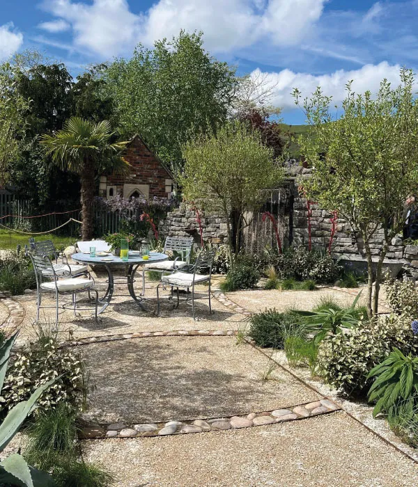 Outdoor garden patio with round metal table and four chairs under blue sky with clouds.