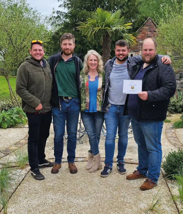 Five people standing on a garden path with greenery behind, one holding a certificate.