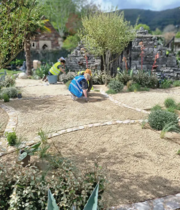 Two people in safety vests working on landscaping a gravel garden with plants and stone pathways.