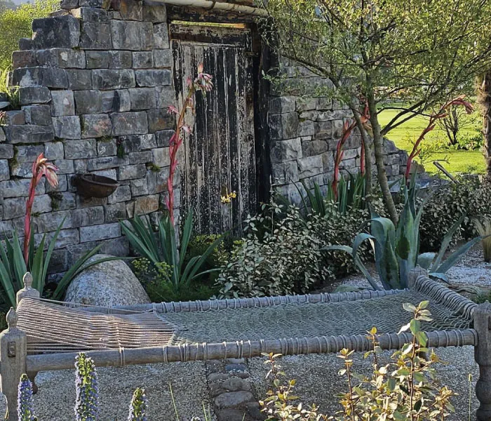 Woven outdoor bench in front of a rustic stone wall with a weathered wooden door surrounded by plants.