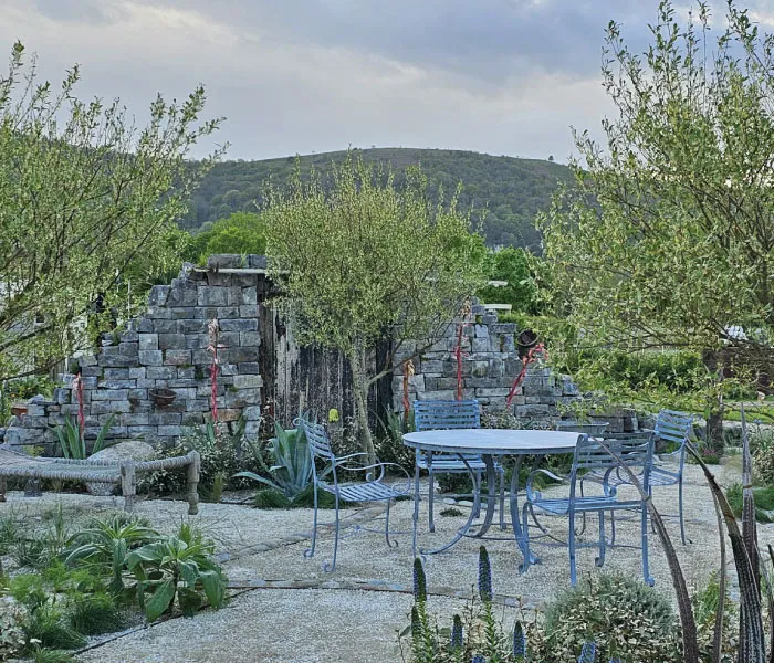 Outdoor garden seating area with metal table and four chairs surrounded by greenery and a stone waterfall wall.