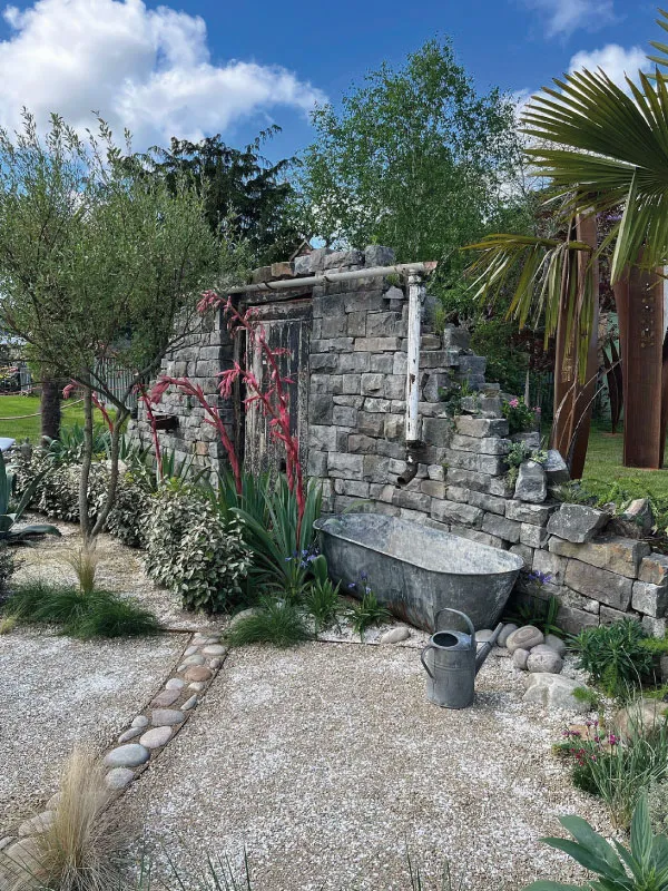 Stone garden wall with old metal bathtub, watering can, and lush green plants under a blue sky.