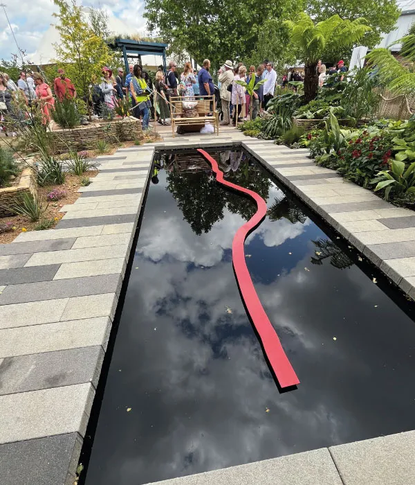 Crowd of people gathered around a reflective rectangular pond with a red curved sculpture in a garden setting.