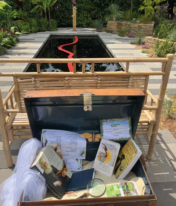 Open wooden box with books, a magnifying glass, and papers on a bench near a rectangular reflective pond.