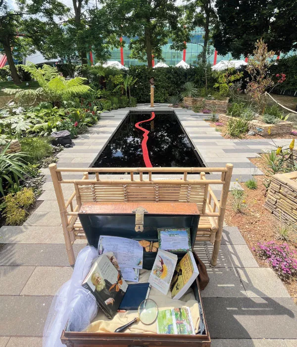 A garden scene showing a bamboo bench facing a reflective water feature with a red zigzag line, surrounded by greenery.