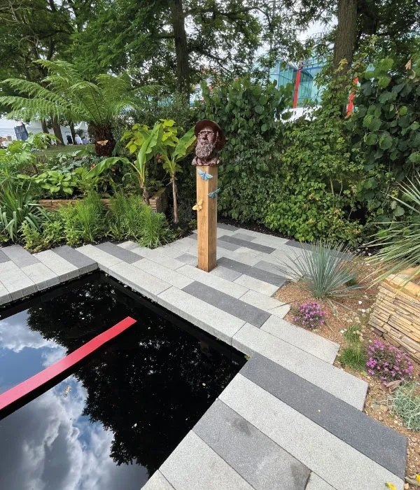 Garden with a wooden pedestal featuring a bearded bust sculpture surrounded by plants beside a reflecting pool.