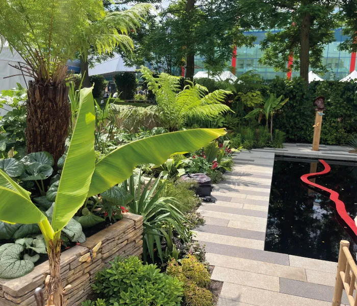 Outdoor garden with diverse green plants and ferns next to a reflective pool with a red ribbon sculpture.
