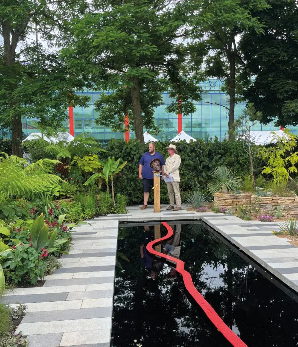 Two men standing by a wooden post with a bust sculpture in a lush garden with a reflective water feature and a red ribbon.