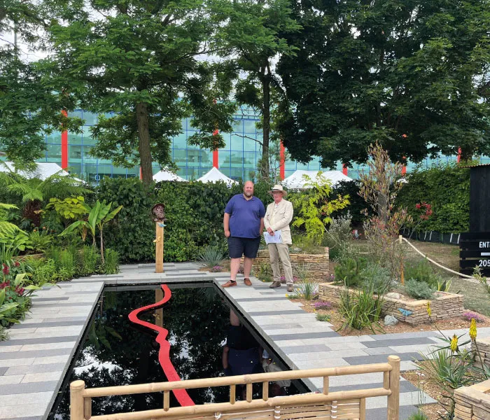 Two men standing beside a rectangular reflecting pool with a red zigzag line, surrounded by trees and plants.