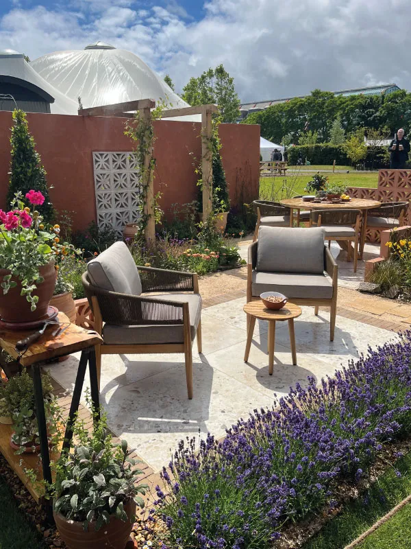 Outdoor patio with two gray cushioned chairs, small wooden table, and vibrant purple flowers in garden.