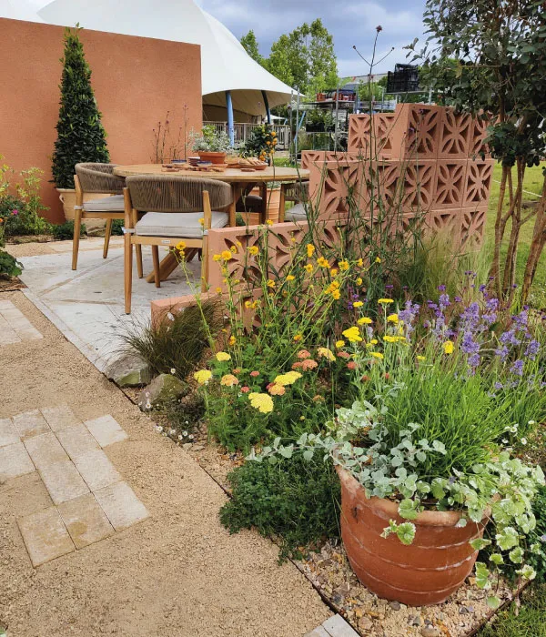 Outdoor dining area with wooden table, cushioned chairs, terracotta decorative blocks, and colorful flowers in a garden.
