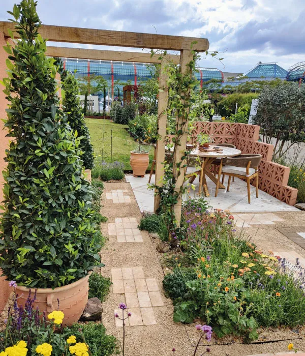 Garden pathway with potted topiary and colorful flowers, leading to a wooden pergola and outdoor seating area.
