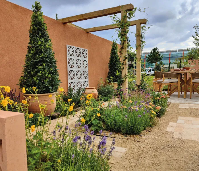 Outdoor garden with terracotta wall, potted topiary, flowering plants, wooden pergola, and patio dining set.