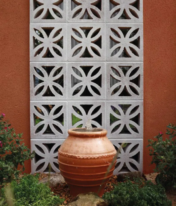 Terracotta pot water fountain in front of a decorative concrete block wall with leafy plants around.