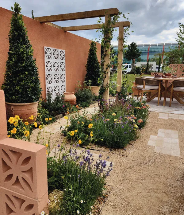 Garden with potted topiary, flowering plants, terracotta wall, wooden pergola, and outdoor seating area.