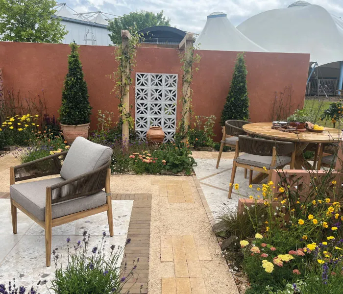 Outdoor patio with wooden chairs, round table, terracotta wall, climbing plants, and colorful flowers.