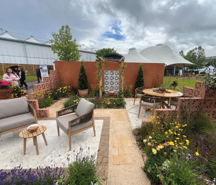 Outdoor seating area with modern chairs, round table, vibrant flowers, and terracotta walls under a cloudy sky.