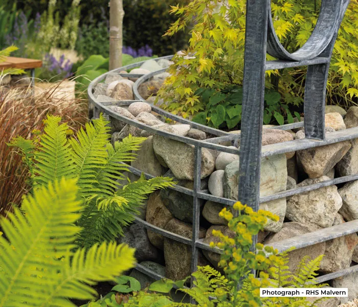 Metal garden structure filled with large stones and surrounded by green ferns and plants.