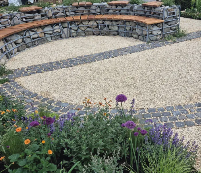 Curved stone bench with wooden seats surrounds a gravel area bordered by cobblestone and colorful flower beds.