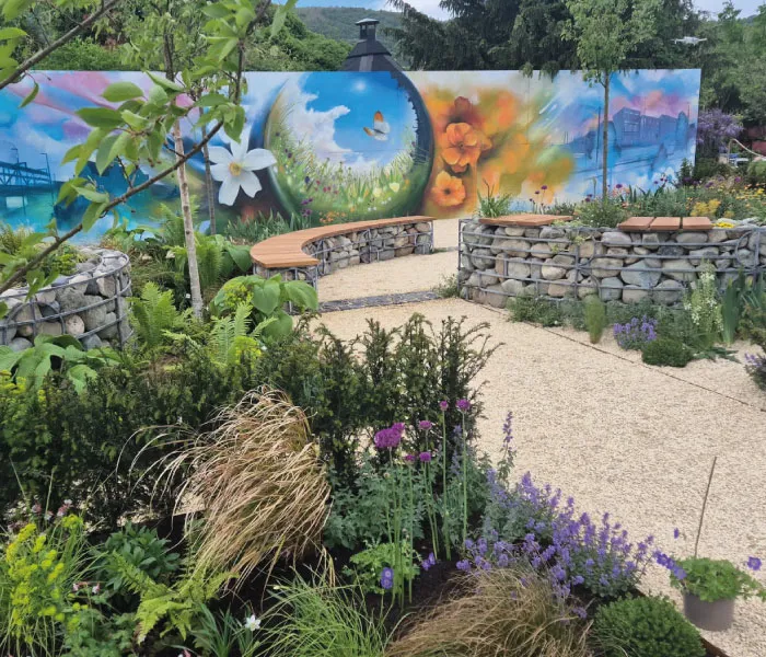 Outdoor garden with stone raised beds, curved wooden benches, lush plants, and a colorful mural with flowers and a butterfly.