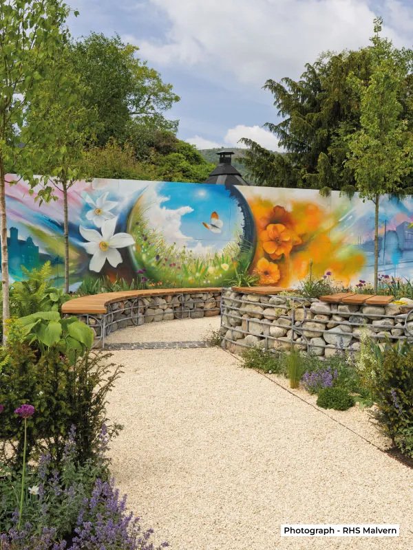Outdoor garden area with stone benches, flowering plants, and a colorful mural of flowers and butterfly on the wall.