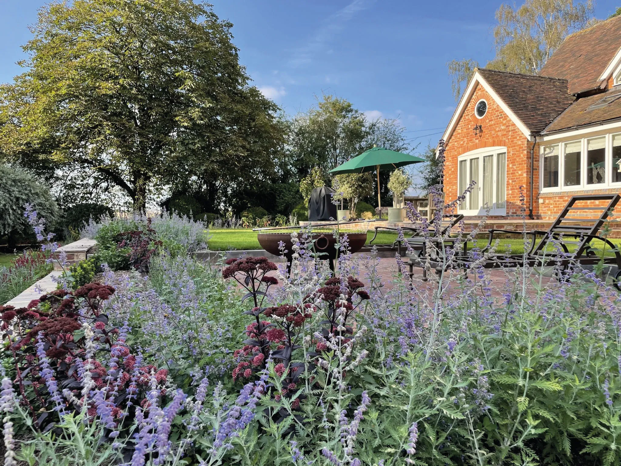 Garden with purple and maroon flowers, green lawn, patio chairs, a fire pit, and a brick house under a clear blue sky.