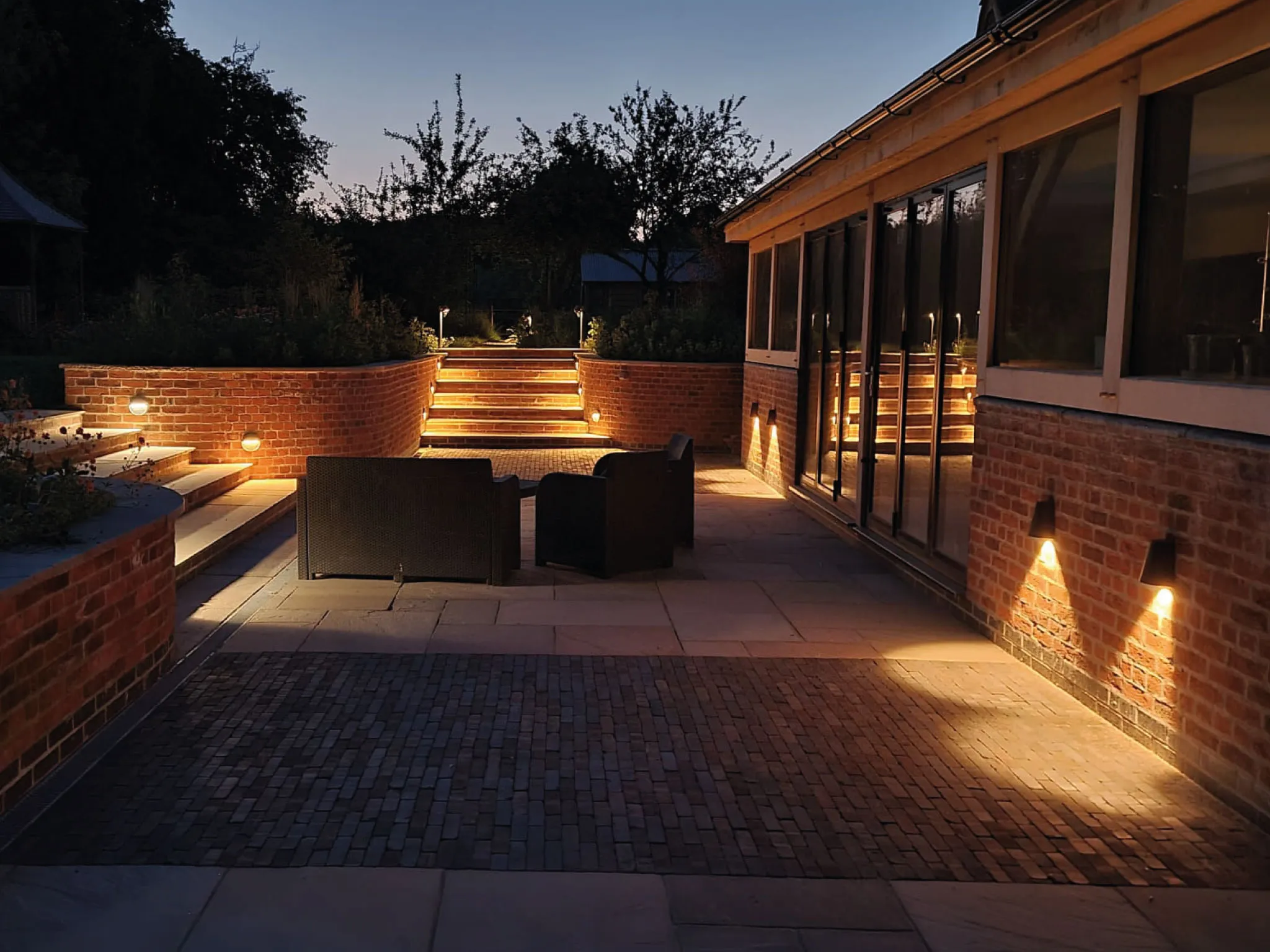 Outdoor patio at dusk with illuminated brick stairs, wall lights, and two dark wicker chairs.