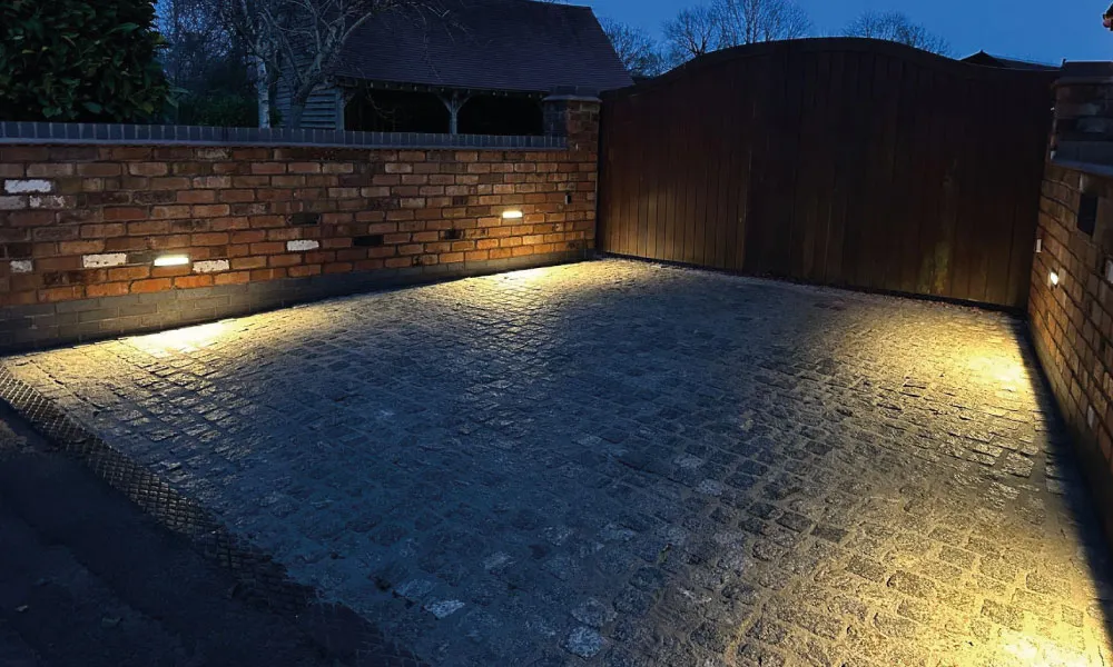 Evening view of a cobblestone driveway with warm ground lights along a brick wall and closed wooden gate.