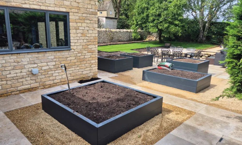 Modern garden with raised black planter boxes filled with soil, patio seating, and a stone house wall.