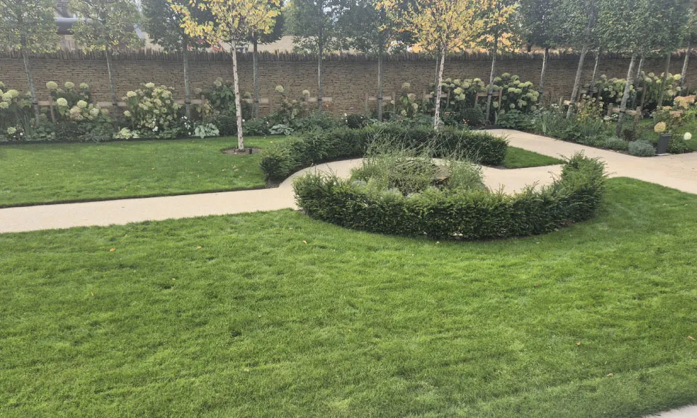 Manicured garden lawn with trees, circular shrub bed, beige walking paths, and hydrangea flowers along a stone wall.