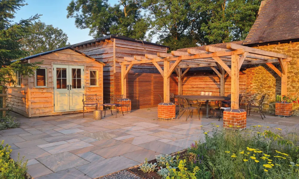 Wooden garden shed and pergola with outdoor seating on stone patio surrounded by greenery and flowers.