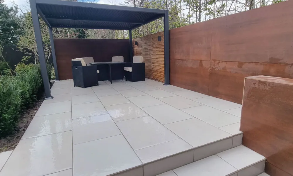 Modern garden patio with light gray tiles, black metal pergola, cushioned chairs, and wooden privacy fencing.