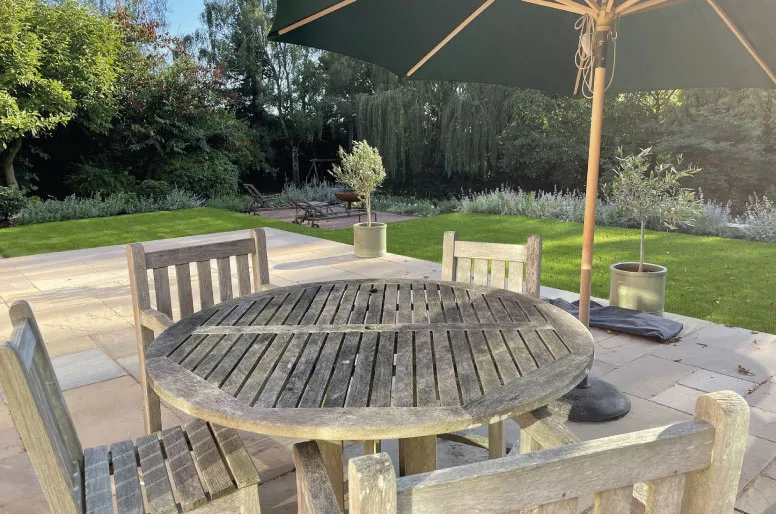 Wooden round garden table with four chairs under a green patio umbrella on stone tile patio.