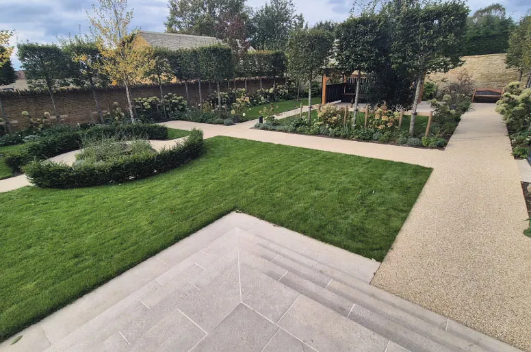 Well-kept garden with green lawn, circular and rectangular hedge beds, and gravel pathways leading to a seating area.