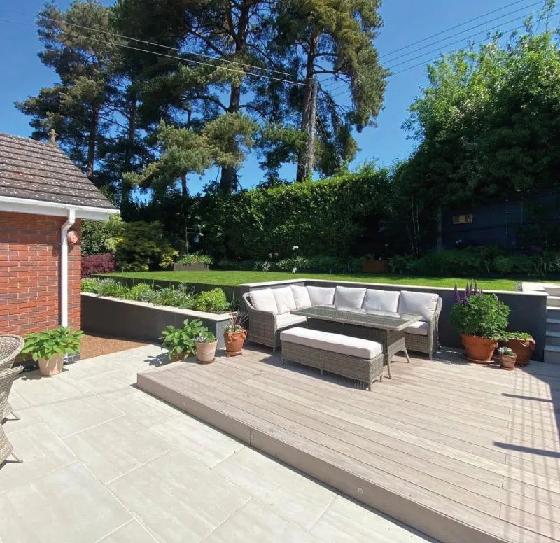 Outdoor patio lounge area with light gray cushioned sectional sofa, glass table, potted plants, and greenery.