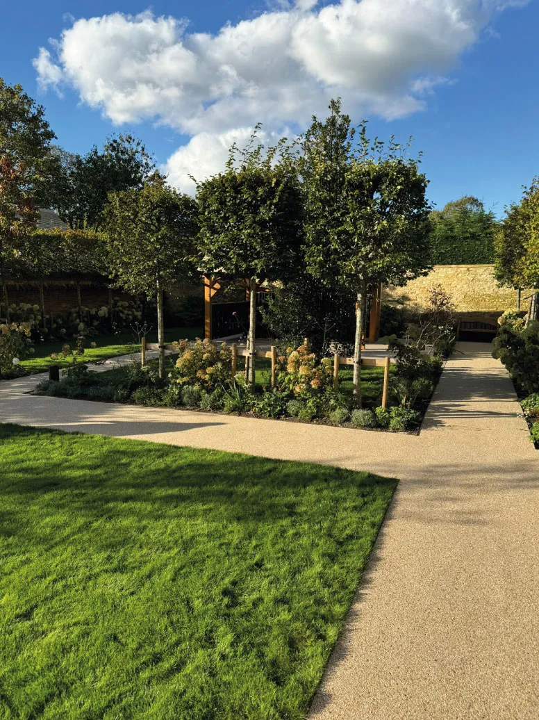 Sunlit garden with trimmed trees, bushes, green lawn, and paved pathways under a partly cloudy sky.