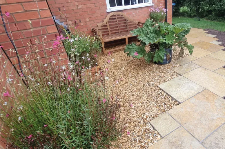 Outdoor patio with beige stone tiles, gravel area, wooden bench, and flowering plants against a brick wall.