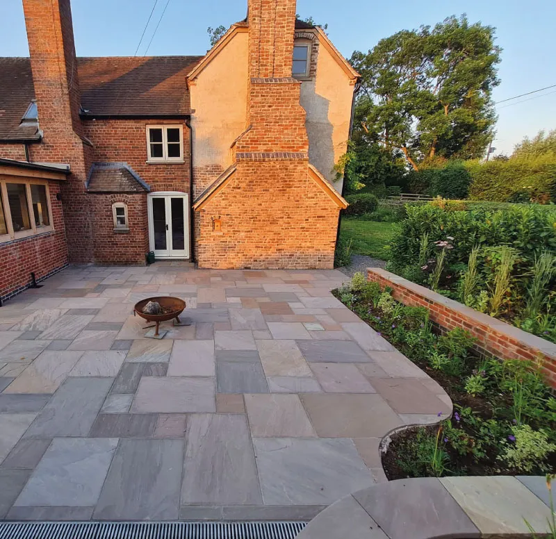 Patio with multicolored stone tiles, metal fire pit, and raised garden bed next to a brick house.